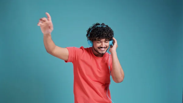 若い笑顔の幸せな陽気な学生インドの男性20代のヘッドフォンで音楽を聴くダンスは 青のスタジオの背景にあるサンゴのTシャツで楽しいです 人々のライフスタイルコンセプト — ストック写真