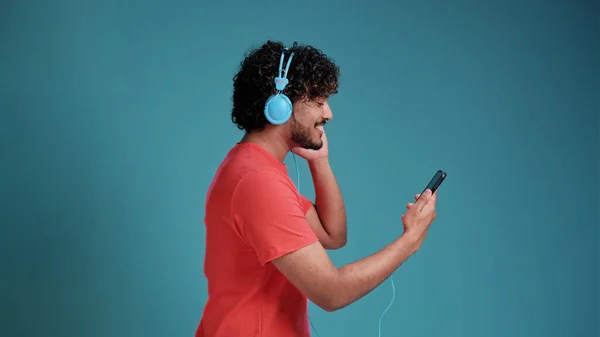明るくハンサムな若いですインディアン男でカジュアルビデオでスマートフォン 新しいヘッドセットを使用して 携帯電話を見て サンゴのTシャツで青のスタジオの背景 コピースペース — ストック写真