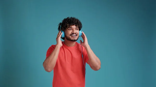 若い笑顔幸せな陽気なインドの男性20代のヘッドフォンで音楽を聴くと 青のスタジオの背景にあるサンゴのTシャツで楽しいです 人々のライフスタイルコンセプト — ストック写真