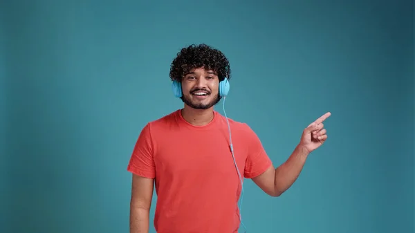 青いスタジオの背景にあるサンゴのTシャツでヘッドフォンを使用して音楽を聴くインド人男性 側に手で非常に幸せなポインティング — ストック写真