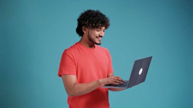 Sakallı, mercan tişörtlü, dizüstü bilgisayarda mail yazan ya da sosyal ağda sohbet eden neşeli Hintli adamın yan görünümü. Stüdyo çekimi mavi stüdyo arka planında izole edildi