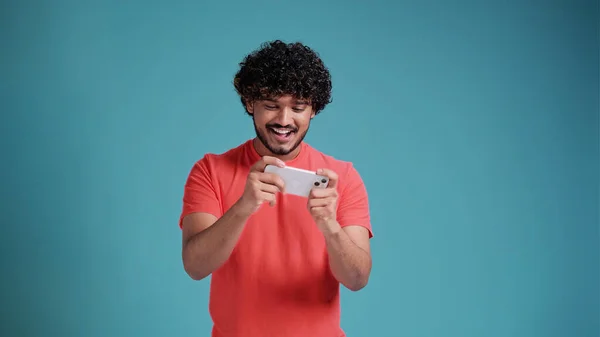 スマートフォンを使用して興奮した幸せな若いインド人男性ゲーマープレーヤー モバイルゲームアプリでゲームをプレイしたり 青いスタジオの背景に隔離された携帯電話を保持するデジタルビデオストリームを見て — ストック写真