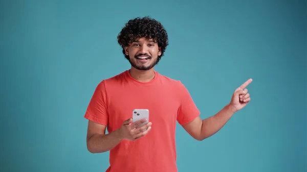 stock image Young indian man using smartphone standing very happy pointing with hand and finger to the side, in coral t-shirt on blue studio background.