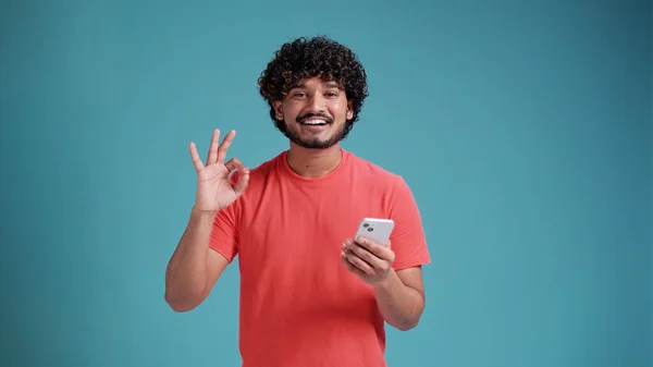 青のスタジオの背景にサンゴのTシャツの優れたシンボル 指でOkサインを行うスマートフォンを使用して若いインド人男性 — ストック写真
