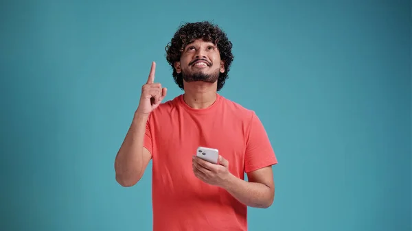 青のスタジオの背景にサンゴのTシャツで 手と指が現れると非常に幸せなポインティングスマートフォンを使用して若いインド人男性 — ストック写真