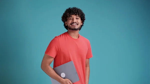 陽気なアラビア語やインド人のひげを生やして若い男は カジュアルなシャツを着て ノートパソコンを持ち 側を見て 喜んで青のスタジオの背景にサンゴのTシャツに笑みを浮かべて — ストック写真