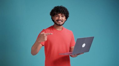 Basit sarı tişörtlü neşeli 20 'li yaşlardaki genç Hintli adam mavi stüdyo arka planında mercan tişörtü ve stüdyo portresi ile dizüstü bilgisayarda iş noktası işaret parmağı kullanıyor. Halk yaşam tarzı konsepti.