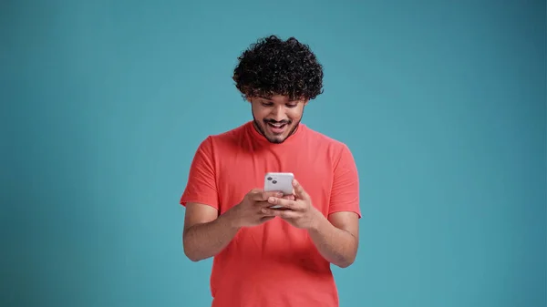 stock image Dreamful marvelous fun young bearded Indian man 20s years old hold in hand use mobile cell phone typing browsing chatting send sms in coral t-shirt on blue studio background studio portrait