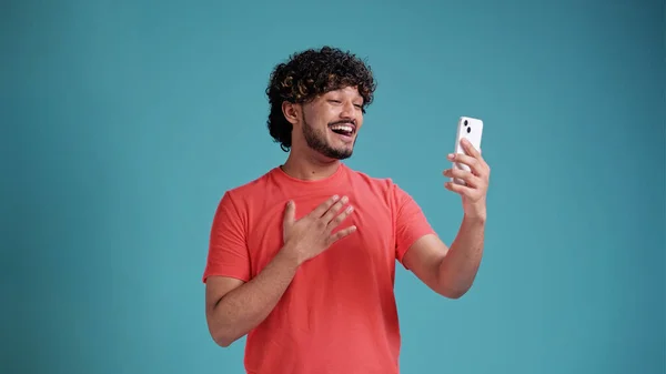 若いですハンサムな笑顔インド人男性作るビデオ通話でスマートフォン 手を振って 青のスタジオの背景に隔離された携帯電話を介してチャット — ストック写真
