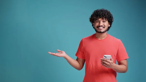 青のスタジオの背景にサンゴのTシャツに手と指で側面に非常に幸せなポインティングスマートフォンを使用して若いインド人の男 — ストック写真