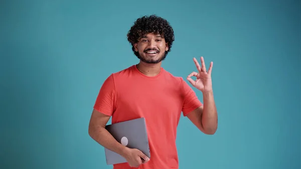 積極的なハンサムなインド人やアラビア人の巻き毛の男 フリーランス ノートパソコンを保持し ポイントOkayジェスチャー 孤立した青のスタジオの背景に立って 優しい笑顔 モックアップ コピースペース — ストック写真