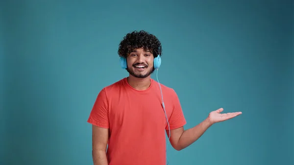 青のスタジオの背景にあるサンゴのTシャツにヘッドフォンで音楽を聴いているラテン系スペイン人男性 手を横に向けて — ストック写真