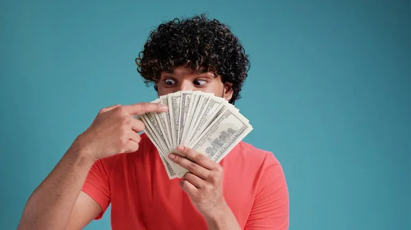 Smiling cheerful happy young bearded latin spanish man 20s years old showing fan of cash money in dollar banknotes looking camera isolated on plain blue background studio portrait.