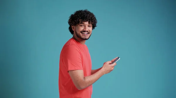 スマートフォンを手にしたラテン系スペイン人男性がカメラを見て笑顔でカジュアル姿で青いスタジオを背景に — ストック写真