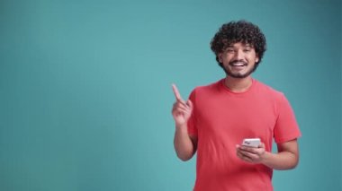 Akıllı telefon kullanan genç latin adam mavi stüdyo arka planında el ve parmak ile çok mutlu bir şekilde duruyor.