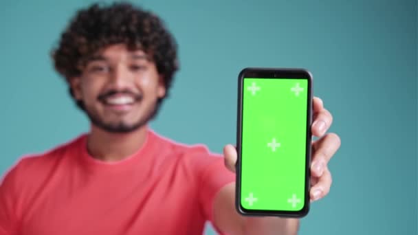 若いラテン人が携帯電話を持っています緑の画面 青のスタジオの背景に空白のクロマキー画面を示しています — ストック動画