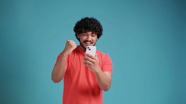 携帯電話を使用して幸せなラテン語やアラビア語の男の勝者スマートフォンを保持興奮民族の男は青のスタジオの背景にサンゴのTシャツで成功を祝う 携帯電話で勝つ賭けに驚いています — ストック写真