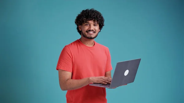 Mavi arka planda mercan tişörtlü, elinde laptop olan genç, yakışıklı Arap ya da Latin bir adam kameraya bakar ve gülümser.