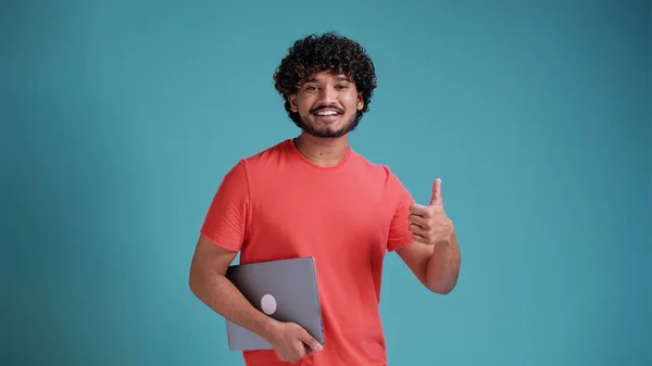 若いハンサムなアラビア語やラテン語の男は 彼の手にノートパソコンと青の背景にサンゴのTシャツに親指を表示カメラを見て笑顔 — ストック写真