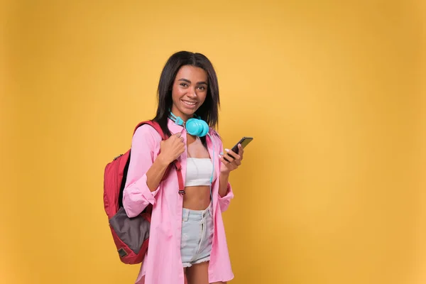 Afro-Amerikalı bir bayan akıllı telefon kullanıyor. İzole edilmiş sarı arka plan üzerinde duruyor. Mutlu bir yüzü var ve gülümsüyor.