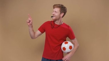 Futbol topu ve düdüğü olan erkek koç bej arka plandaki boş bir yeri işaret ediyor.