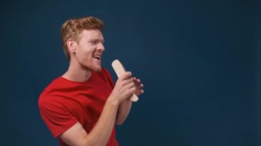 Kızıl saçlı, genç ve çekici bir adam koyu mavi bir arka planda çalılara şarkı söylüyor.