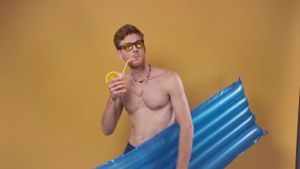 红头发的男人穿着海滩短裤 有一个充气床垫 鸡尾酒把他的手指指向侧面 黄色背景的暑假概念 — 图库视频影像