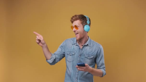 在手机应用程序上听音乐的年轻人 带着耳机和舞步站在空旷的空间上 与黄色背景隔离 — 图库视频影像