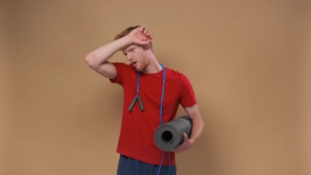 健身和生活方式的概念 照片中英俊的中年男子 做完健身锻炼后疲倦了 拿着跳绳和瑜伽垫 擦去额头上的汗珠 背景是米色的 — 图库视频影像