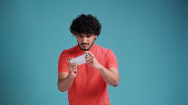 Akıllı telefon kullanan, mobil video oyunu uygulamasında oyun oynayan ya da mavi stüdyo arka planında izole edilmiş dijital video akışını izleyen heyecanlı, genç esmer erkek oyuncu.