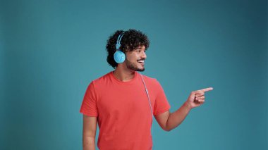 Esmer adam müzik dinliyor. Mavi stüdyo arka planında mercan tişörtlü kulaklık takıyor. Yan yana çok mutlu bir şekilde işaret ediyor.