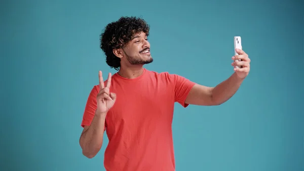 Genç, yakışıklı, esmer adam akıllı telefondan video görüşmesi yapıyor, cep telefonuyla el sallıyor, sohbet ediyor mavi stüdyo arka planında izole edilmiş..