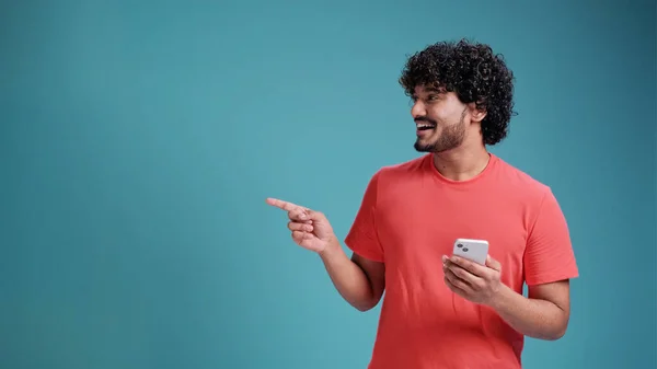 青のスタジオの背景にサンゴのTシャツに手と指で側面に非常に幸せなポインティングスマートフォンを使用して若いSwarthy男 — ストック写真