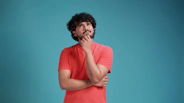 stock image swarthy man in coral t-shirt on blue studio background thinking pondering looking for an idea