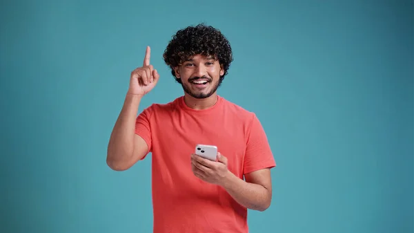 青のスタジオの背景にサンゴのTシャツで 手と指が現れると非常に幸せなポインティングスマートフォンを使用して若いヒスパニックまたはアラブ人の男 ロイヤリティフリーのストック写真