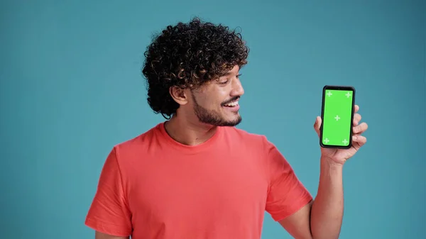 青のスタジオの背景にサンゴのTシャツに空白のクロマキー画面を示し 緑の画面を持つ携帯電話を保持若いヒスパニックまたはアラブ人の男 ロイヤリティフリーのストック画像