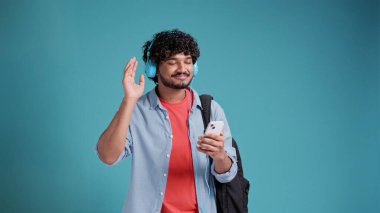 Mavi stüdyo arka planında kulaklıklı ve akıllı telefonlu müzik dinleyen Hintli ya da İspanyol genç öğrenci..