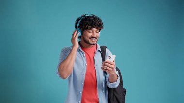 Latin Amerikalı ya da Arap genç bir öğrenci mavi stüdyo arka planında kulaklık ve akıllı telefonla müzik dinliyor..