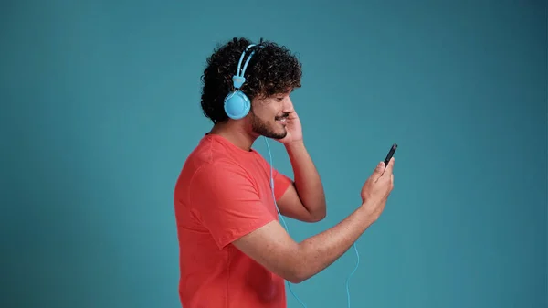 明るくハンサムな若いラテン語やアラビア語の男でカジュアルなビデオを見ますスマートフォン 新しいヘッドセットを使用して 携帯電話を見て 青のスタジオの背景にサンゴのTシャツ コピースペース — ストック写真