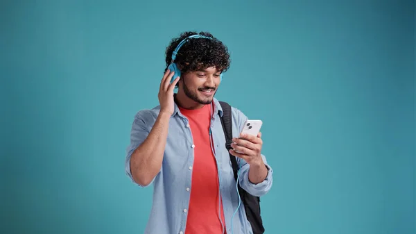 インド人やスペイン人の若い男性学生は 青いスタジオの背景にヘッドフォンやスマートフォンで音楽を聴く — ストック写真