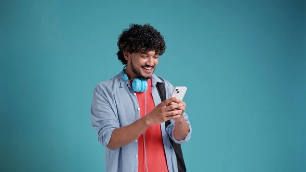 インド人やスペイン人の若い男性でカジュアルとともにヘッドフォンやスマートフォンの使用アプリと笑顔で青のスタジオの背景 — ストック写真