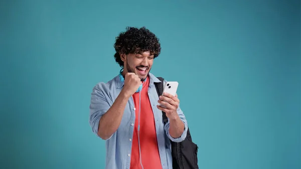 ラティーノかアラブの若い男子学生バックパックを手にスマートフォンで成功と勝利を喜びます 青いスタジオを背景に — ストック写真