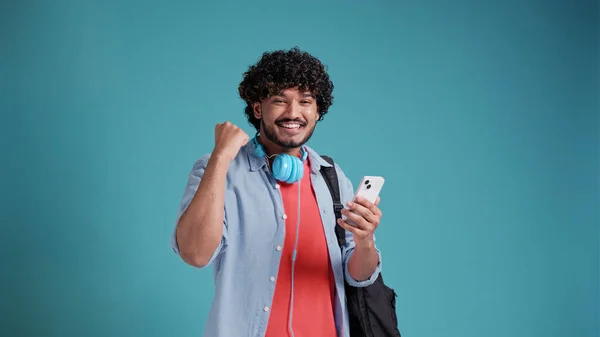 バックパックを持つインド人またはスペイン人の若い男性学生は 彼の手の中にスマートフォンで成功と勝利を喜びます 青いスタジオを背景に — ストック写真