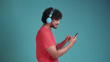 Yakışıklı Hintli genç adam, günlük kıyafetleriyle akıllı telefondan müzik dinliyor, yeni kulaklık takıyor, cep telefonuna bakıyor ve gülümsüyor, mavi stüdyo arka planında mercan tişörtü giyiyor.