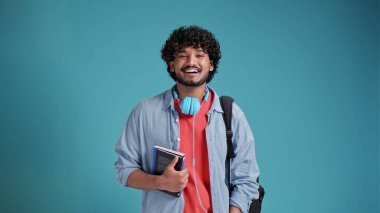 Sırt çantalı çok ırklı erkek öğrenci mavi bir stüdyoda gülümsüyor.