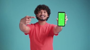 Genç Hintli bir adam elinde yeşil ekranlı bir akıllı telefon tutuyor. Mavi stüdyo arka planında mercan tişörtlü, boş renkli bir ekran gösteriyor.