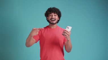 20 'li yaşlarda mutlu, sakallı, etnik kökenli genç bir adam cep telefonu kullanıyor. Akıllı telefonuna işaret ediyor. Mavi stüdyo arka planında tişörtü var.