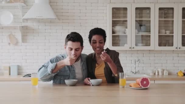 キッチンで朝食を食べるハッピーな多民族カップル キッチンでアフリカ系アメリカ人の若い女性とヨーロッパ人男性がスプーンで美味しい健康的なお粥を食べる — ストック動画