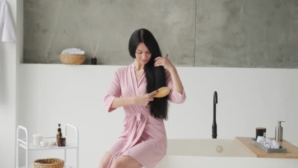 バスルームに座っている間彼女の髪を慰めるバスローブで豪華な髪の美しい若いブルネットの女性 — ストック動画