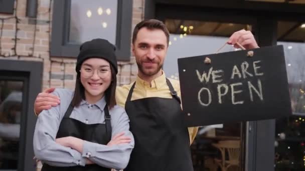 ようこそ オープンしました 2人のフレンドリーなウェイター レストラン カフェ バーの外に立って看板Openを示す白人男性とアジアの女性 — ストック動画